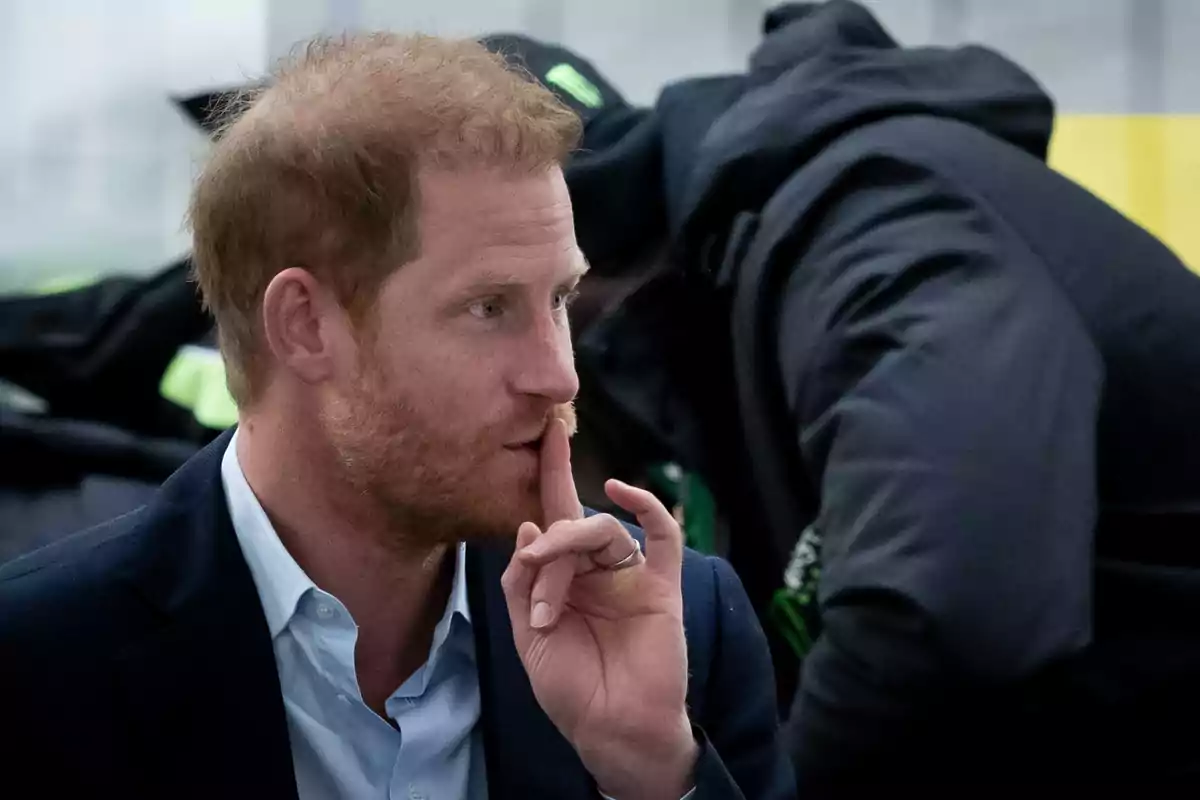 Prince Harry with a beard makes a shushing gesture with his index finger over his lips while wearing a dark suit and a light jersey.