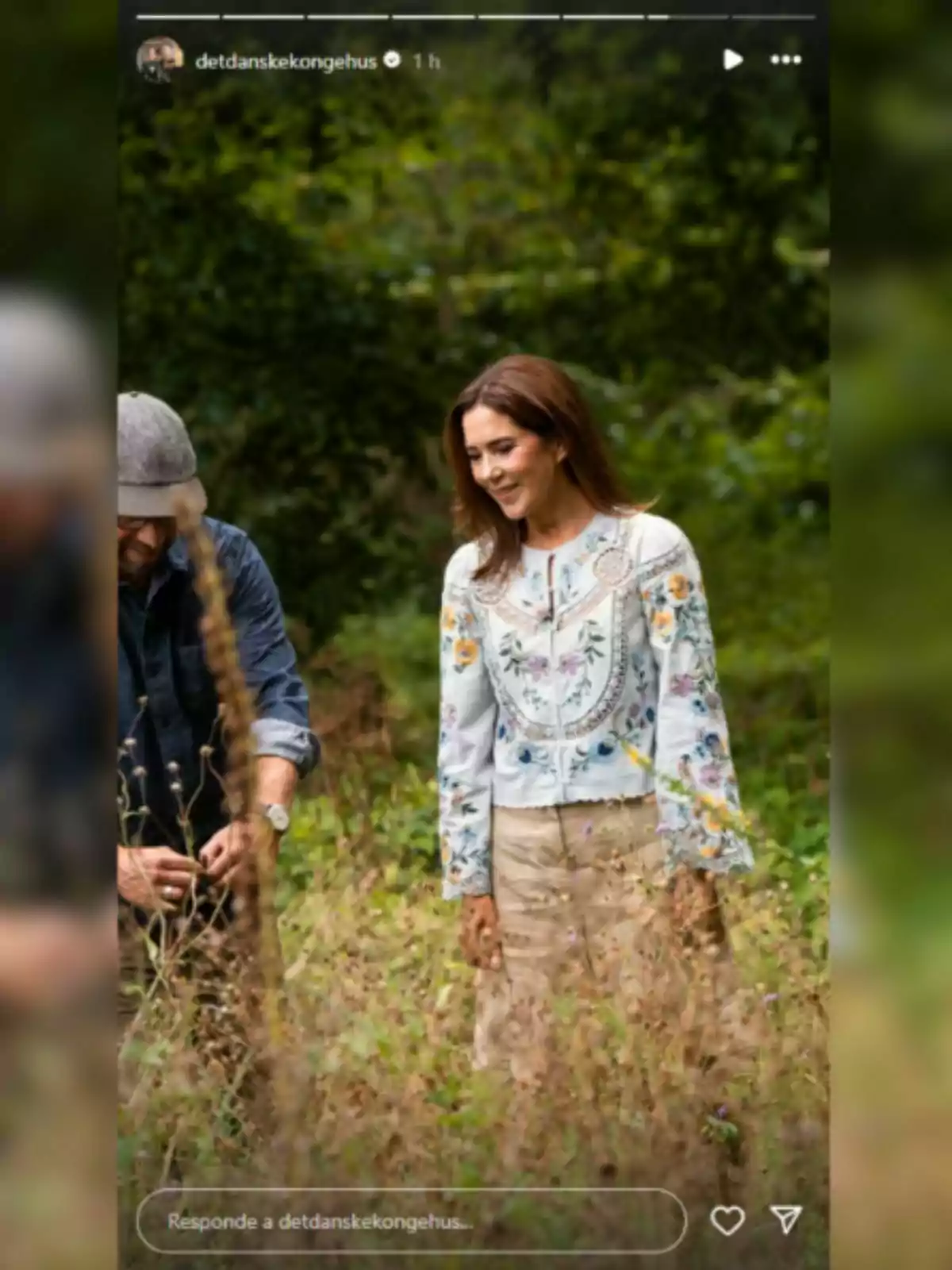 Mary of Denmark, smiling in an embroidered blouse and beige skirt, walking through a field surrounded by greenery next to a partially visible man
