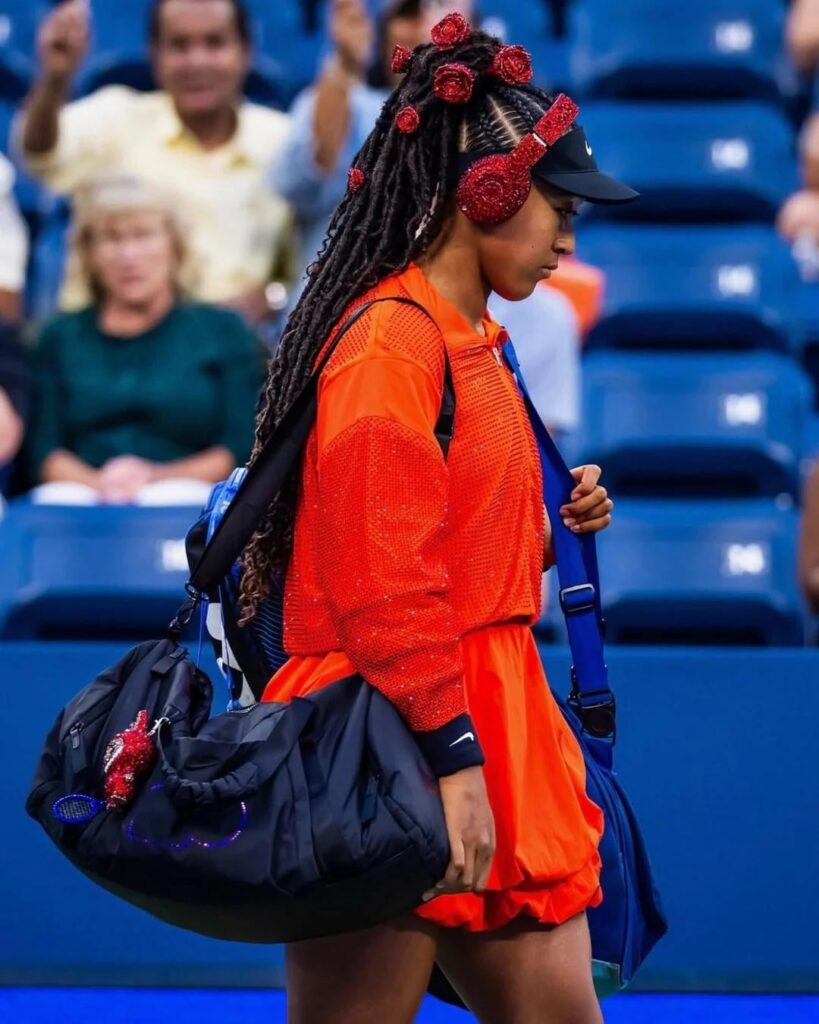 Naomi Osaka served one of the best tennis fits I’ve ever seen: cropped jacket, bubble-hem skirt, crystal roses in her hair, bedazzled headphones, and a matching bedazzled Labubu she’s christened Billie Jean Bling