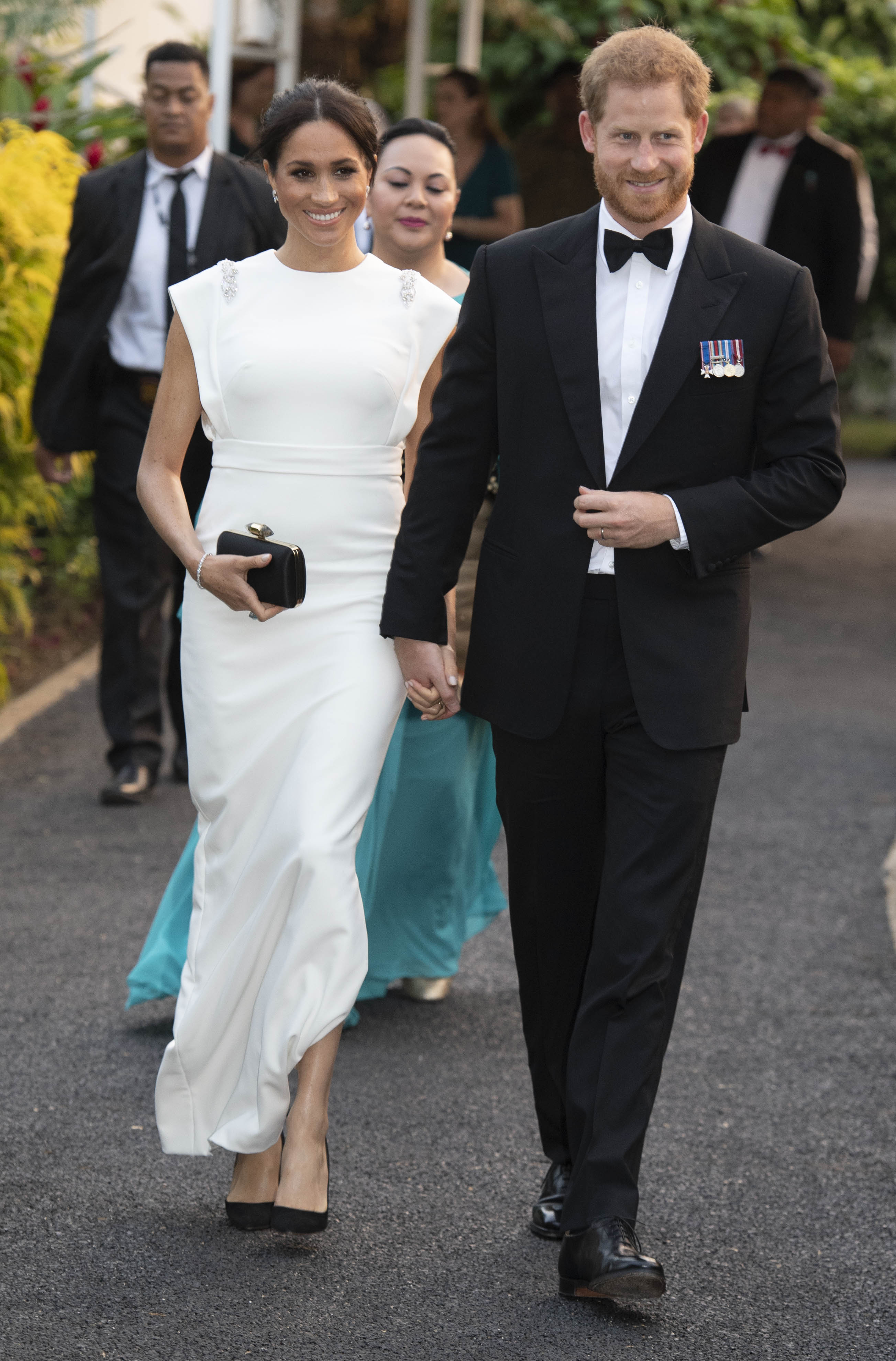 Meghan Markle wearing a white gown walking and holding hands with Prince Harry, wearing a tuxedo