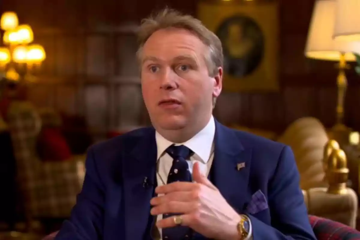 Grant Harrold in a blue suit speaking in an elegantly decorated room with lamps and classic furniture