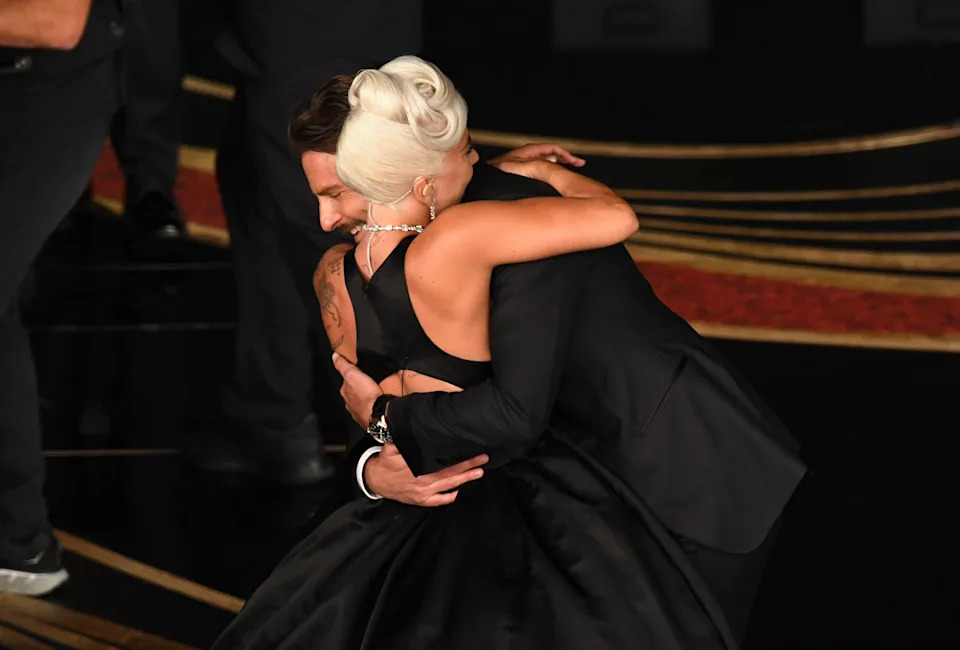 Two people in formal attire share a warm embrace on a red carpet event