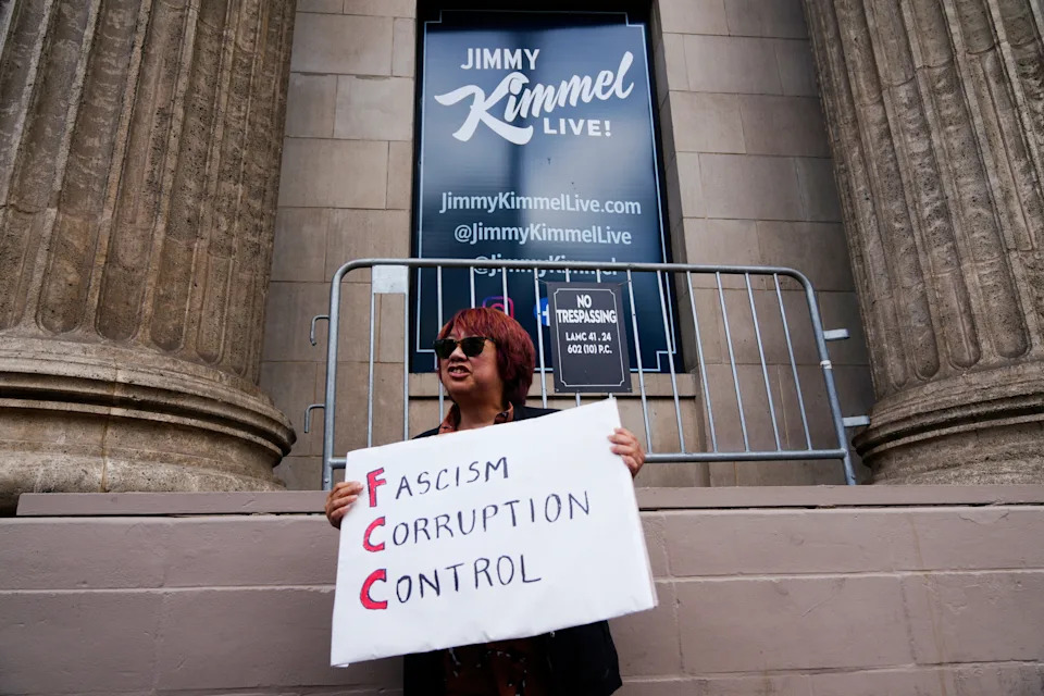 Someone standing near a Jimmy Kimmel Live poster holds a sign with the letters FCC highlighted that reads: Fascism, corruption, control.