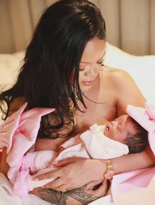 A person holds a newborn wrapped in a soft blanket, smiling gently. They wear an elegant outfit with floral accents on the shoulders