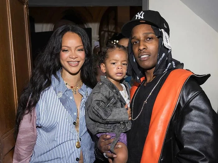 Two celebrities posed together with a child. One wears a mixed-pattern shirt, and the other sports a cap and a jacket with orange detail