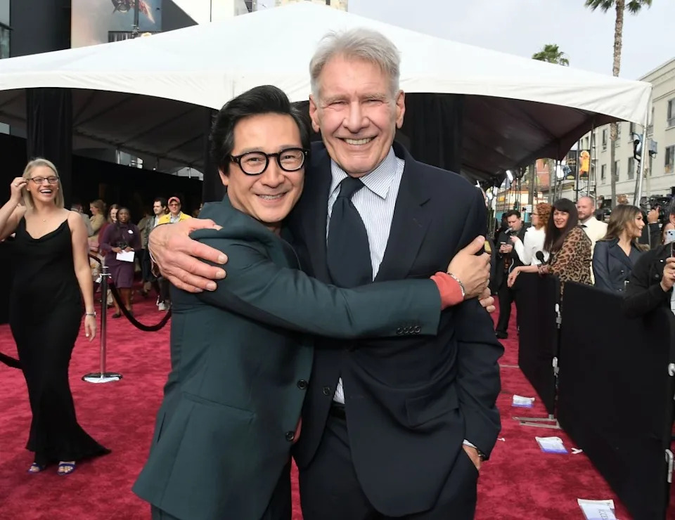 Two men smiling and embracing on a red carpet. A lively event atmosphere surrounds them