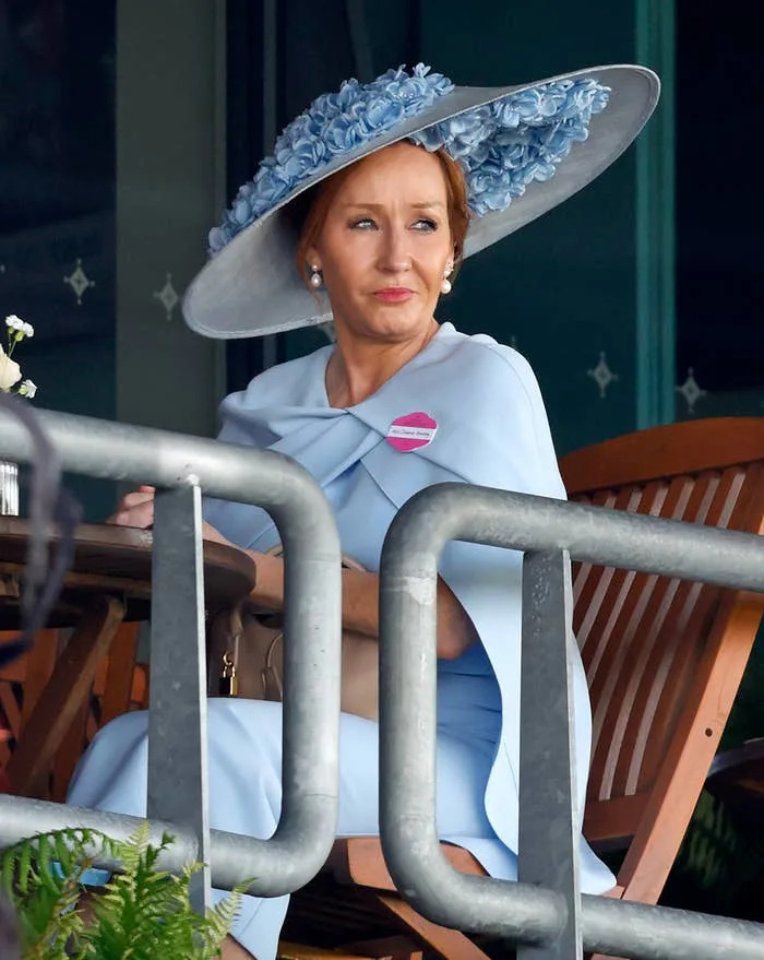 Person seated, wearing an elegant wide-brimmed hat adorned with flowers and a stylish outfit