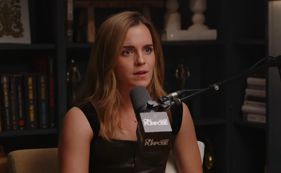 Person in a podcast studio setting, wearing a sleeveless top, speaking into a microphone. Books and decorative items in the background