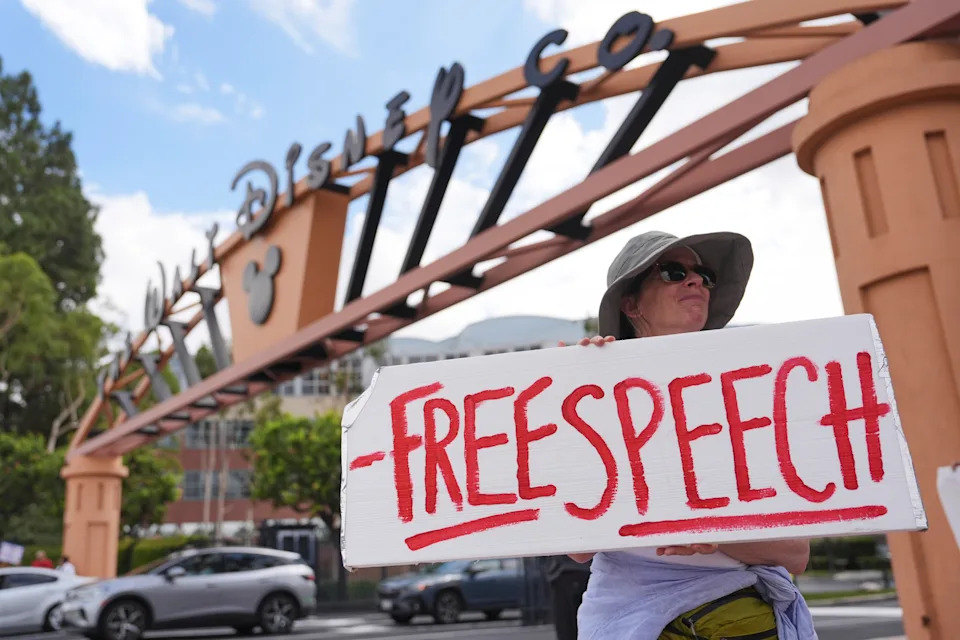 A demonstrator standing below a large corporate sign reading Walt Disney Co. holds a hand-written sign reading: Free speech.