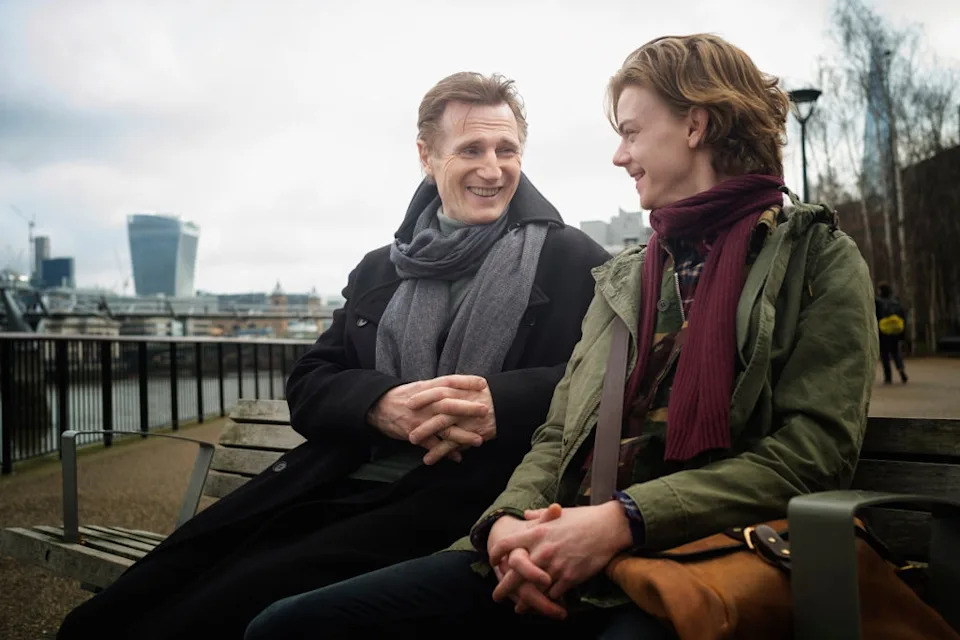 Two people sit on a bench outdoors, engaged in conversation. One wears a long coat and scarf; the other in a jacket and scarf, holding a bag
