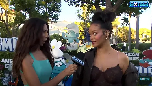 A celebrity in a black lace dress speaks with a reporter on a thematic outdoor set, featuring animated character decorations
