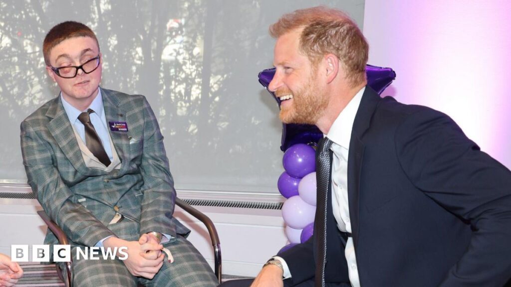 Declan sits beside Prince Harry at the awards ceremony in London. Declan is wearing a blue and grey checked three piece suit, blue shirt and dark tie. Prince Harry is wearing a dark suit, white shirt and dark blue tie.