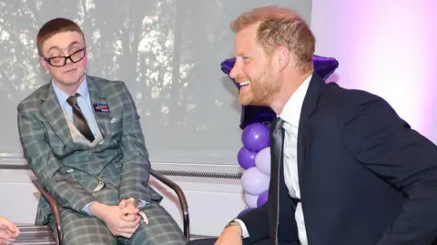 WellChild Awards Declan sits beside Prince Harry at the awards ceremony in London. Declan is wearing a blue and grey checked three piece suit, blue shirt and dark tie. Prince Harry is wearing a dark suit, white shirt and dark blue tie.