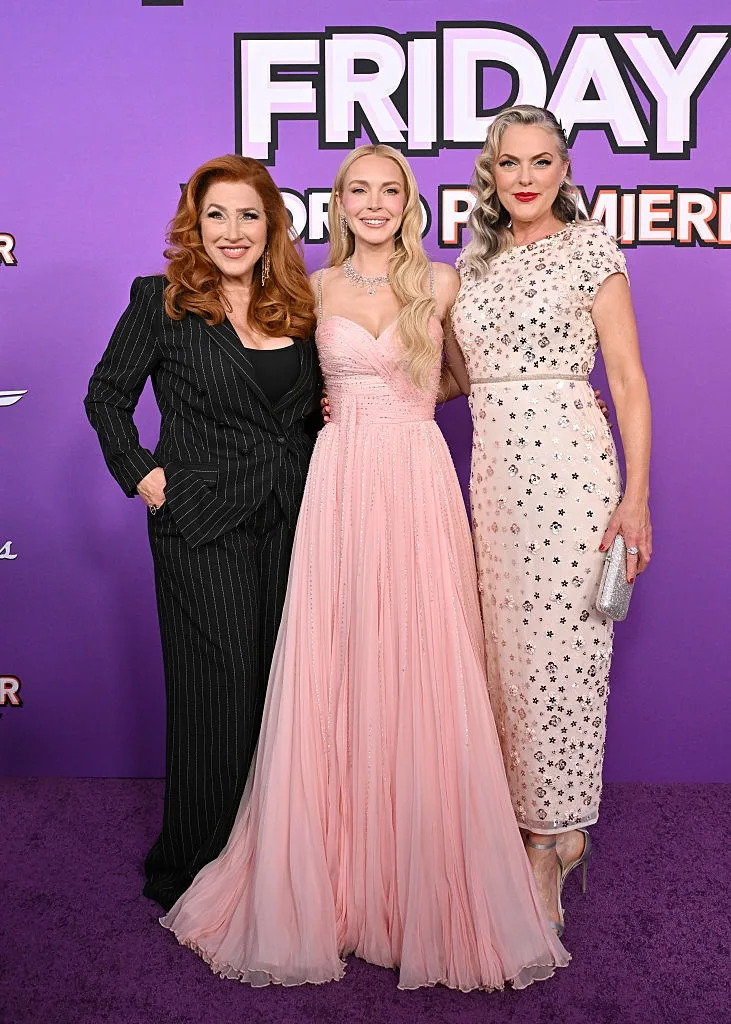 Three women on a red carpet; one in a pinstripe suit, one in a strapless gown, and one in a floral dress, pose at a TV and movie premiere event