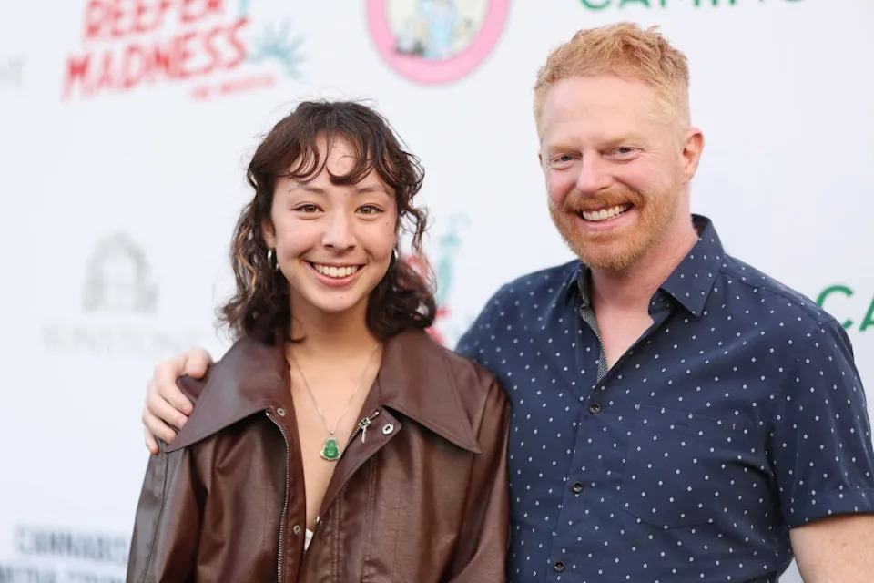 Two people smiling on a red carpet, one wearing a leather jacket and the other a polka-dot shirt, attending a media event