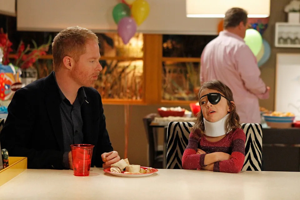 A man and a girl with an eye patch and neck brace sit at a table during a party. The man looks concerned as the girl appears upset