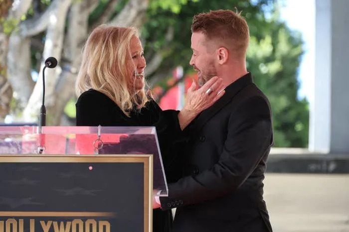Two people share a warm moment on a stage with a Hollywood-themed podium in front of them