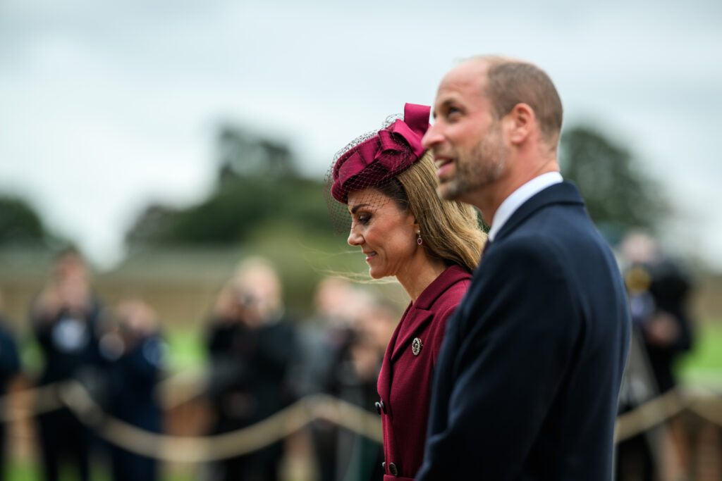 The Prince of Wales smiles as he walks with the Princess of Wales in front of a row of photographers.