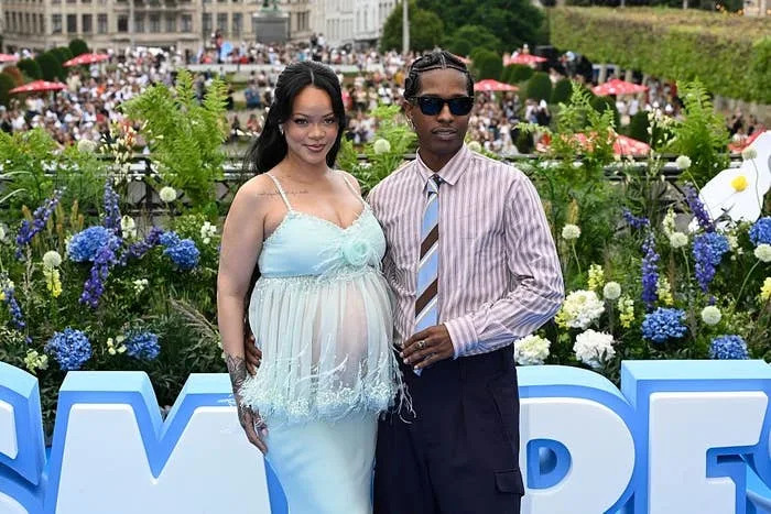 Celebrity couple poses at an outdoor event; she wears a flowing dress, and he wears a striped shirt with a tie