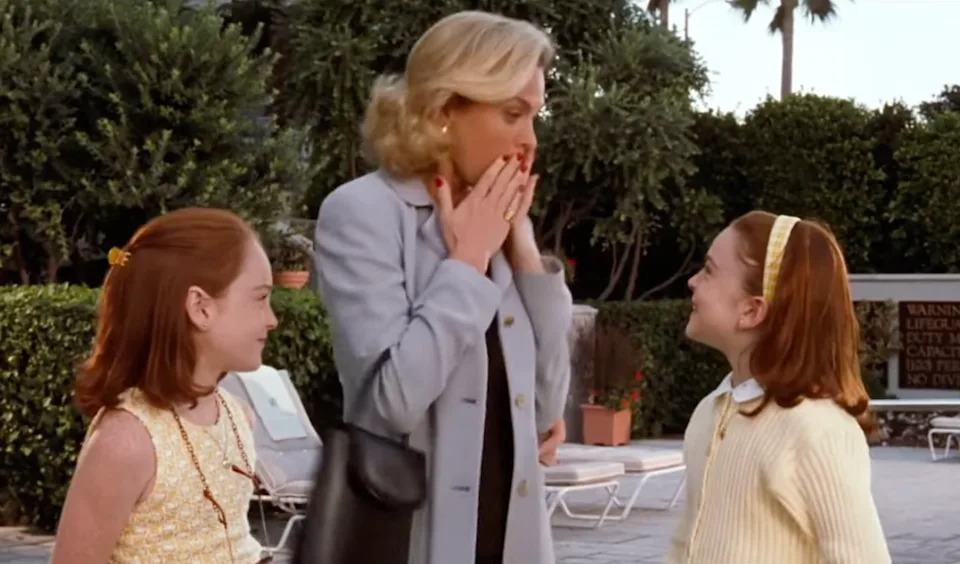A woman in a light coat covers her mouth while talking to two young girls outdoors