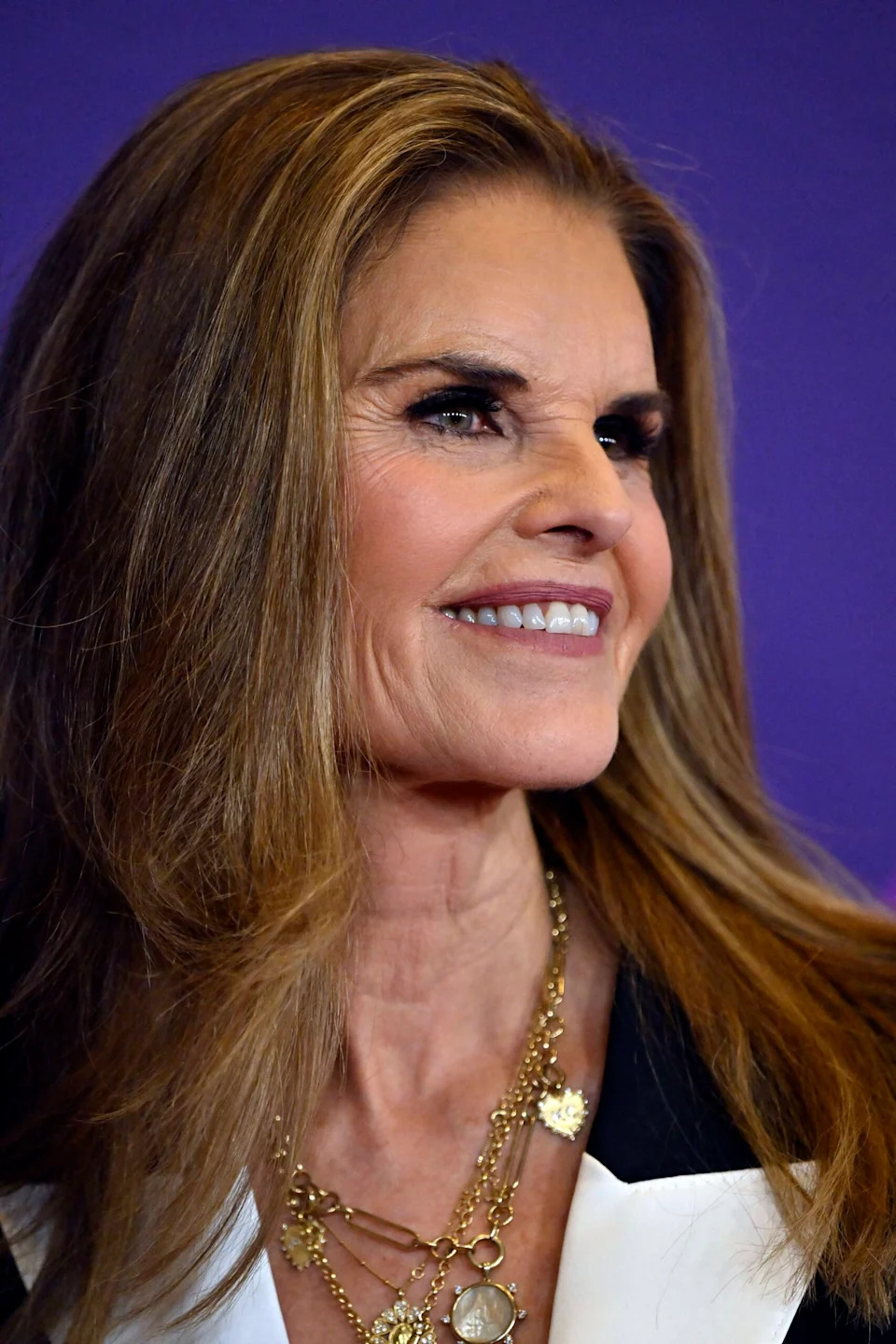 Maria Shriver smiles in a close-up photo, wearing layered gold necklaces and a black-and-white outfit.