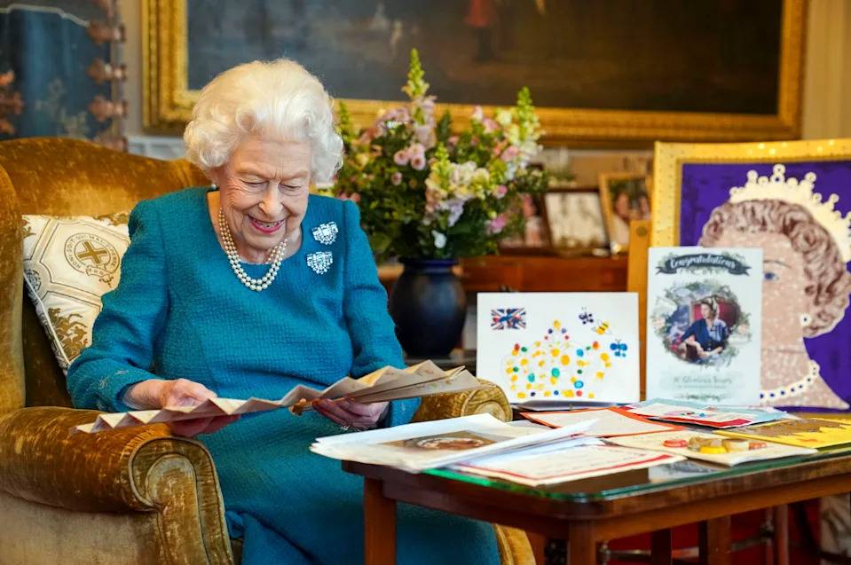 Queen Elizabeth wearing a blue dress smiling and looking at cards and a fan set on a table