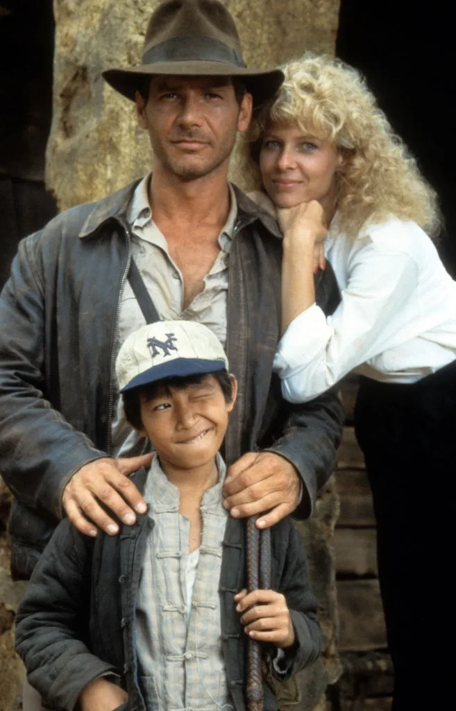 Three people from an adventure film: a man in a fedora, a woman in a blouse, and a child wearing a cap, standing together outdoors