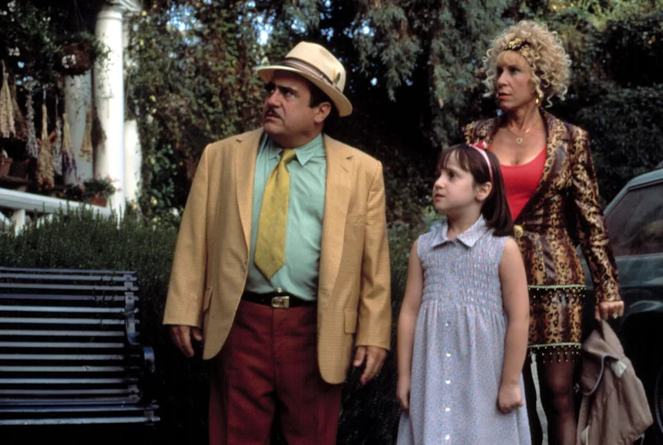 A man in a suit, a woman in a patterned jacket, and a young girl in a dress stand outside looking surprised