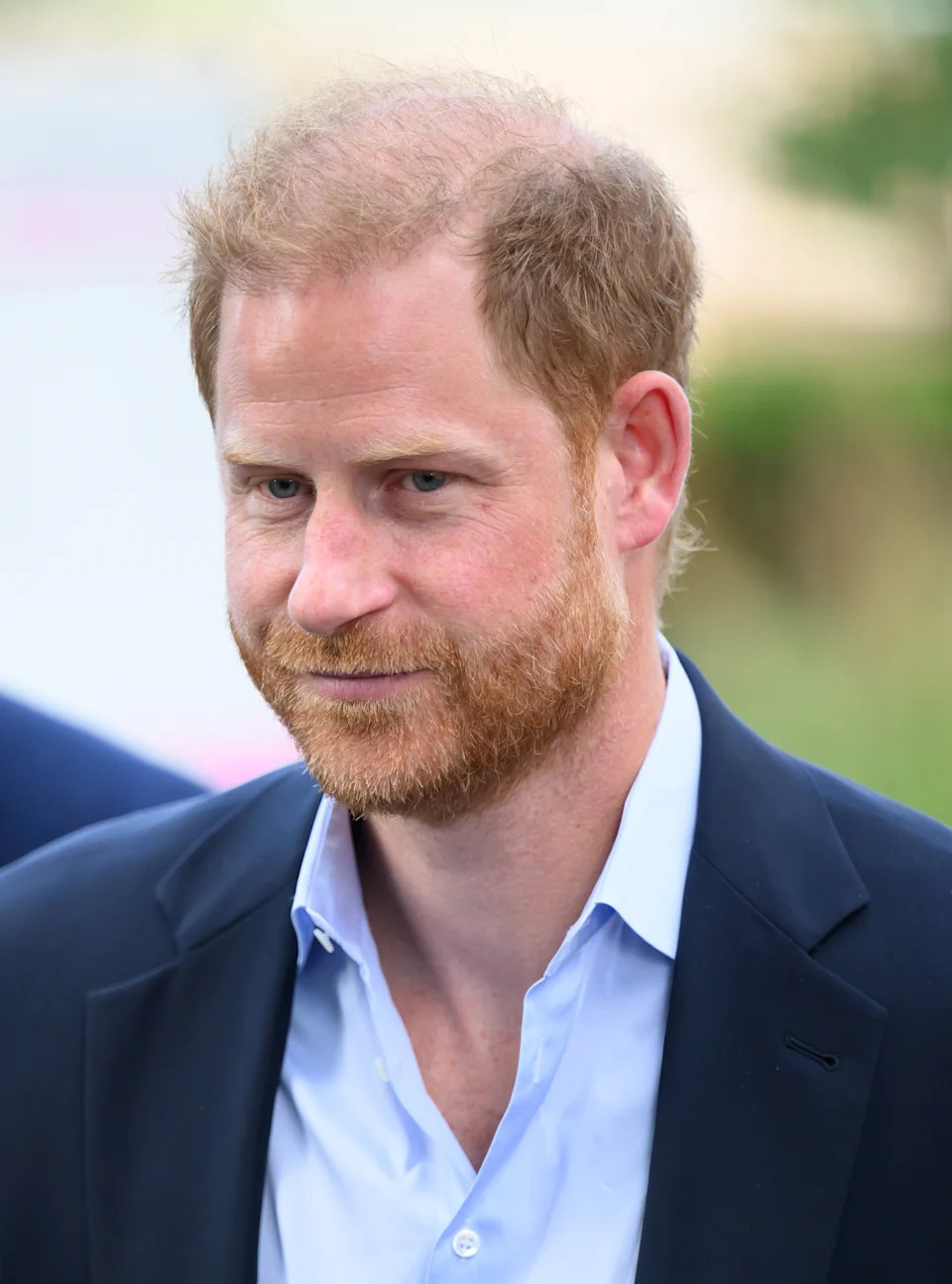 Prince Harry with a short beard and casual suit jacket walks outdoors, looking ahead with a slight smile