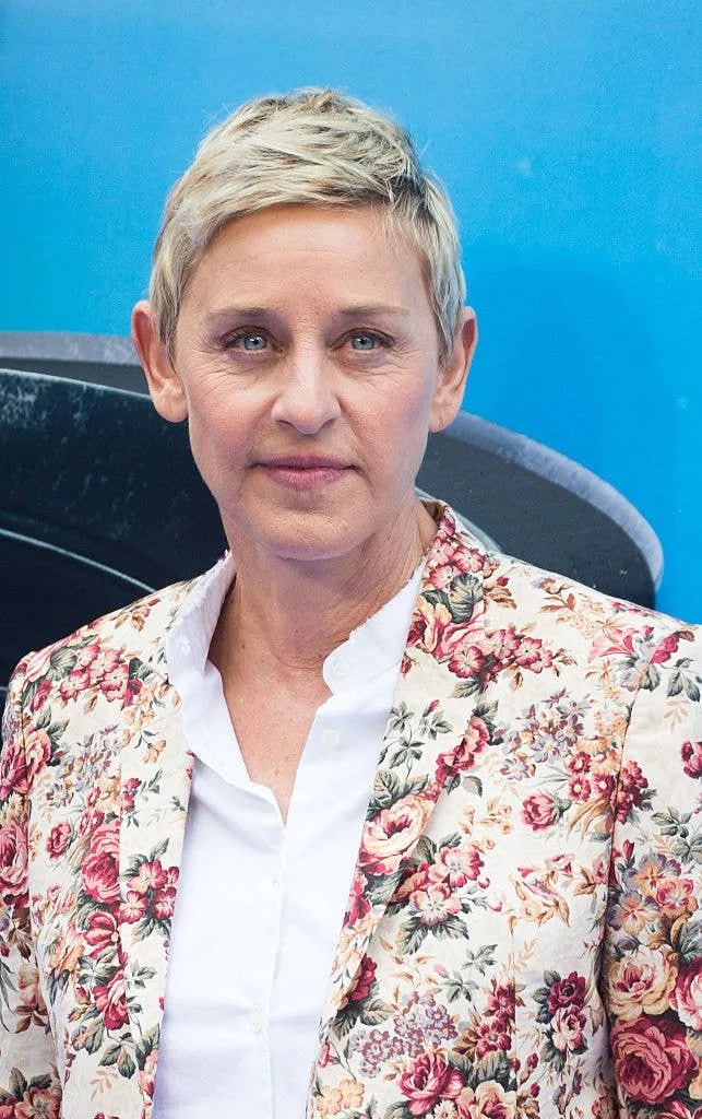 Person with short hair wearing a floral patterned jacket over a white shirt, standing in front of a blue background