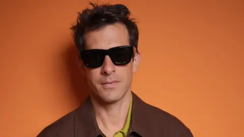 Getty Images Portrait of Mark Ronson, wearing a brown jacket, green shirt and dark glasses in front of an orange backdrop