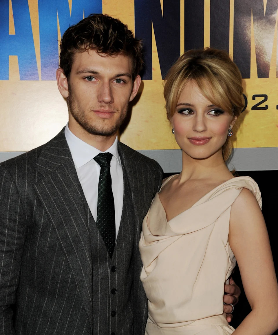 A man and woman pose at a media event. The man wears a pinstripe suit, and the woman wears an elegant, draped dress