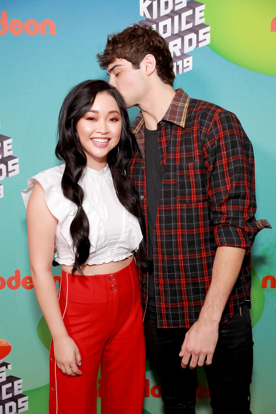 Two individuals pose at a Kids' Choice Awards event. One wears a flared blouse and high-waisted pants, and the other wears a plaid shirt, leaning in for a kiss