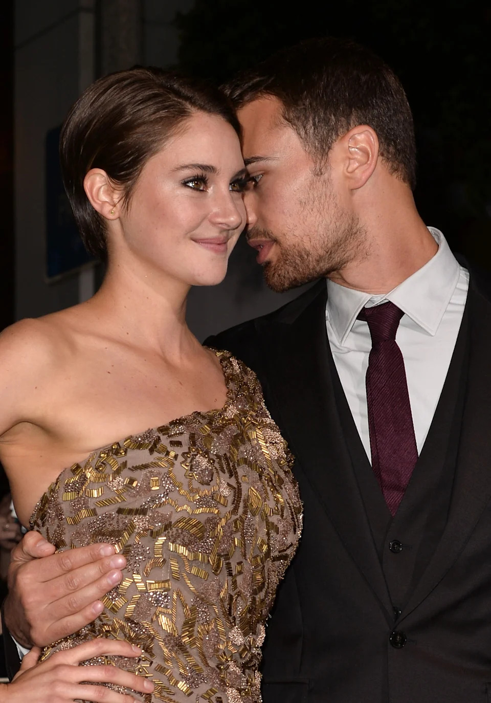 Two people in formal attire pose closely; one in a sequined, one-shoulder dress, the other in a suit with a tie, looking at each other