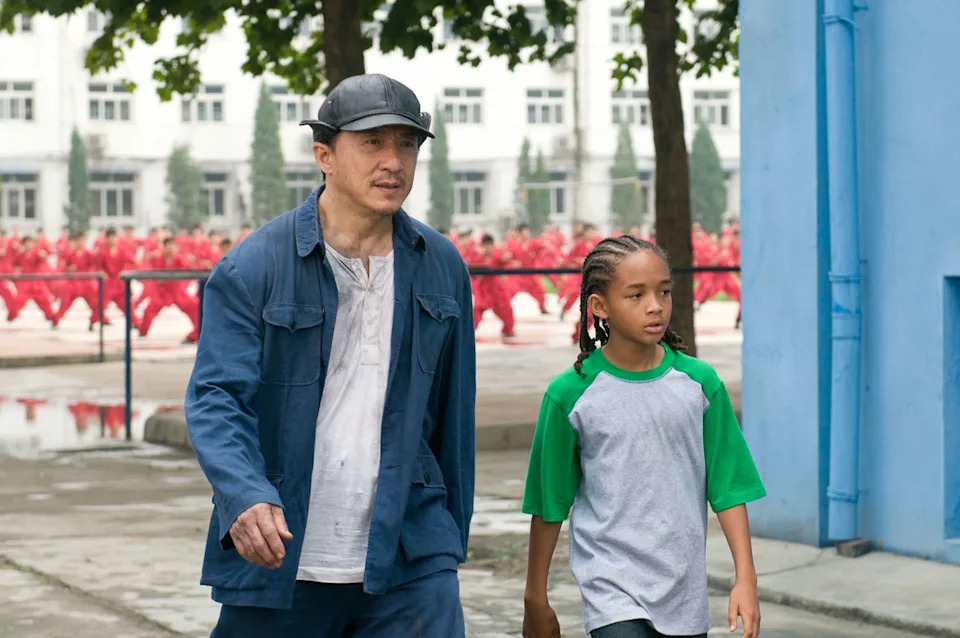 Two individuals walking in a courtyard; one in casual blue attire and cap, the other in a two-tone t-shirt. Background shows a line of red uniforms