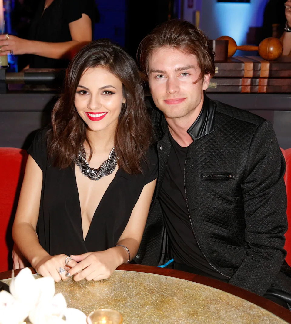 Two people sitting at a table, smiling at the camera. The person on the left wears a V-neck outfit with a statement necklace