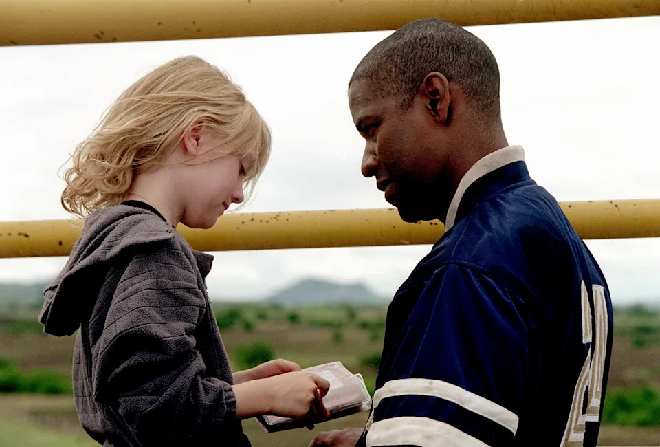 A girl shows a book to a man wearing a varsity jacket. They are outdoors, with a fence and landscape in the background