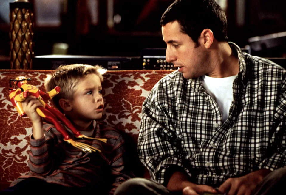A man in a plaid shirt and a young boy with a toy sit on a couch, engaged in conversation, in a scene likely from a TV show or movie
