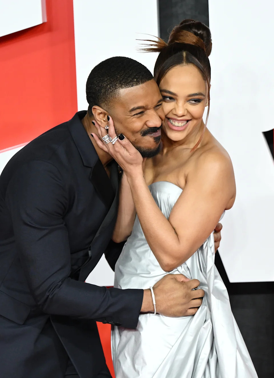 Two people smiling and embracing on a red carpet. One is wearing a sleek dark suit, and the other is in a strapless, elegant gown