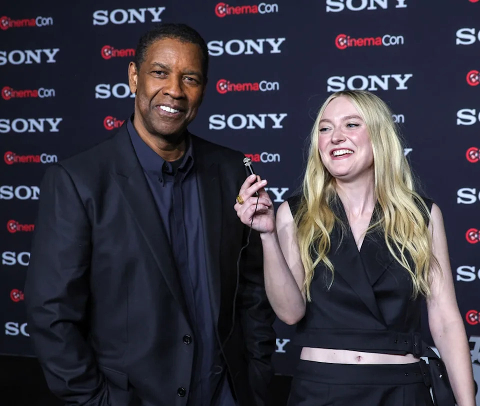 Two people smiling at a CinemaCon event, one in a dark suit, the other in a sleeveless top and pants, holding a microphone