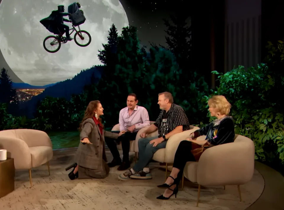 TV show set with a host kneeling, three guests seated, and a backdrop of a moonlit bicycle scene