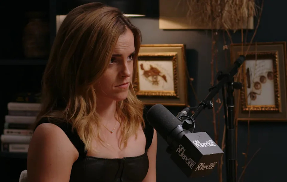 Person with long hair, wearing a black top, sits at a podcast setup with a microphone labeled "Purpose."