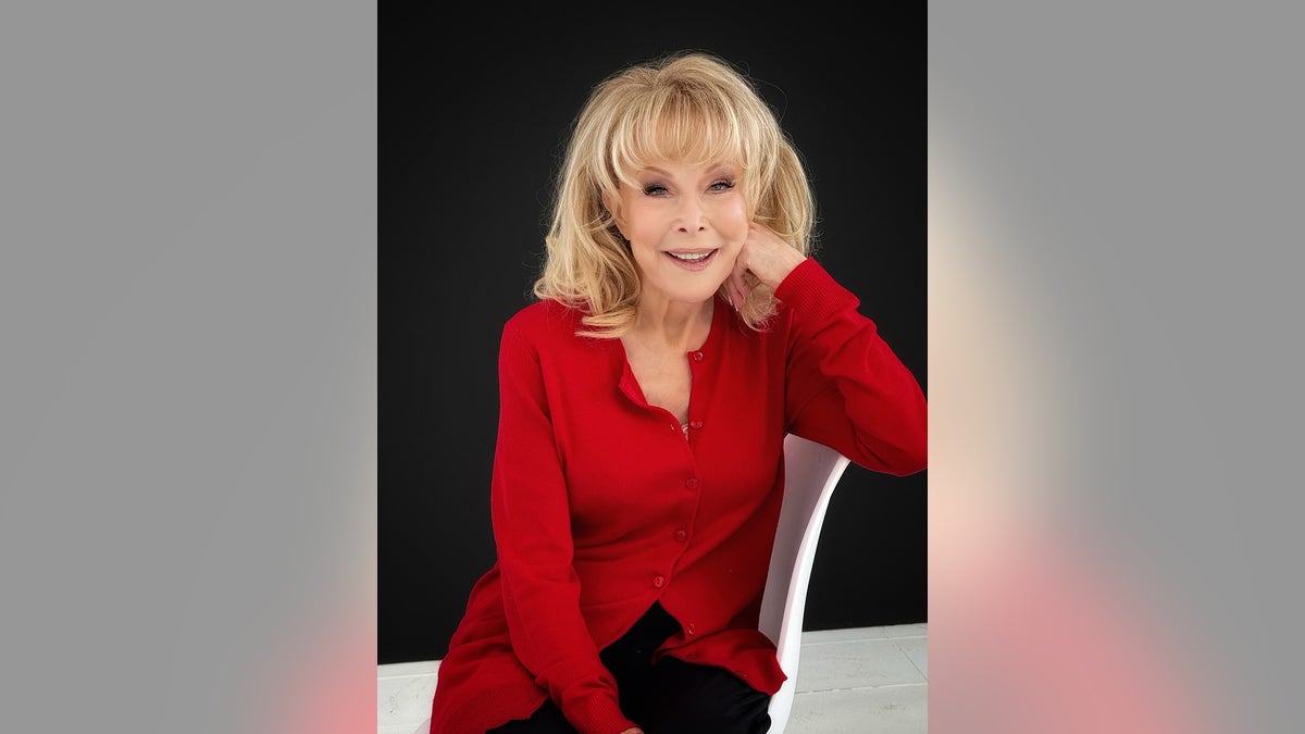 Barbara Eden wearing a red blouse and sitting on a white chair.