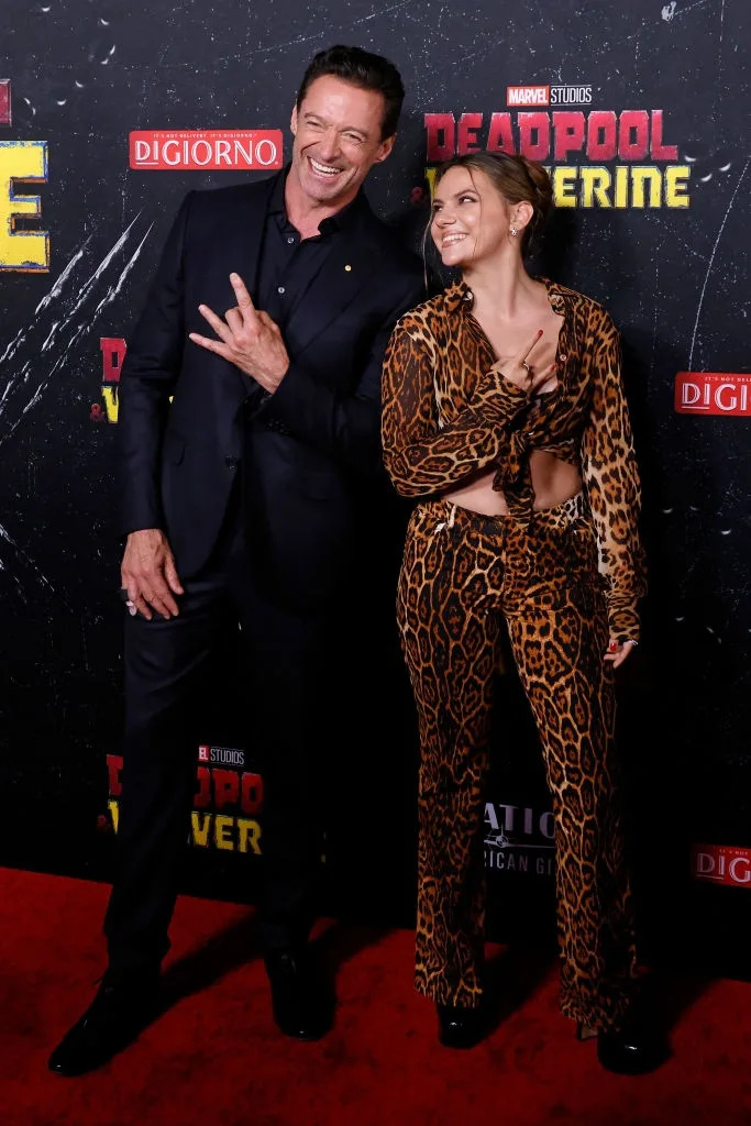 Two individuals smiling on a red carpet at a "Deadpool 3" event. One wears a dark suit, the other wears a leopard print outfit