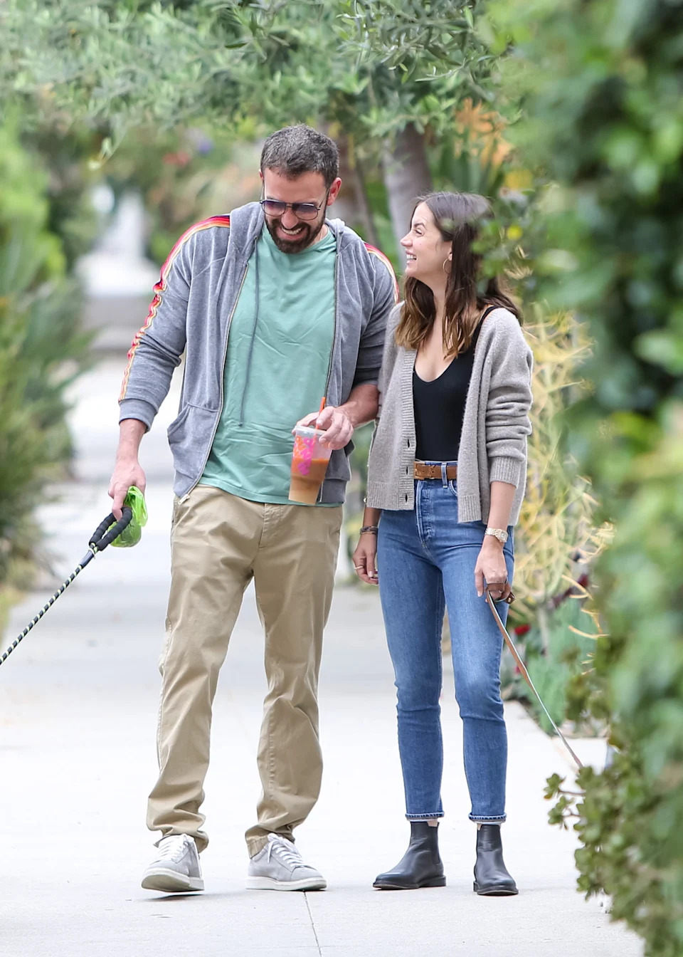 Two people walking dogs on a sidewalk, wearing casual outfits. One wears a hoodie and the other a cardigan and jeans. Both are smiling