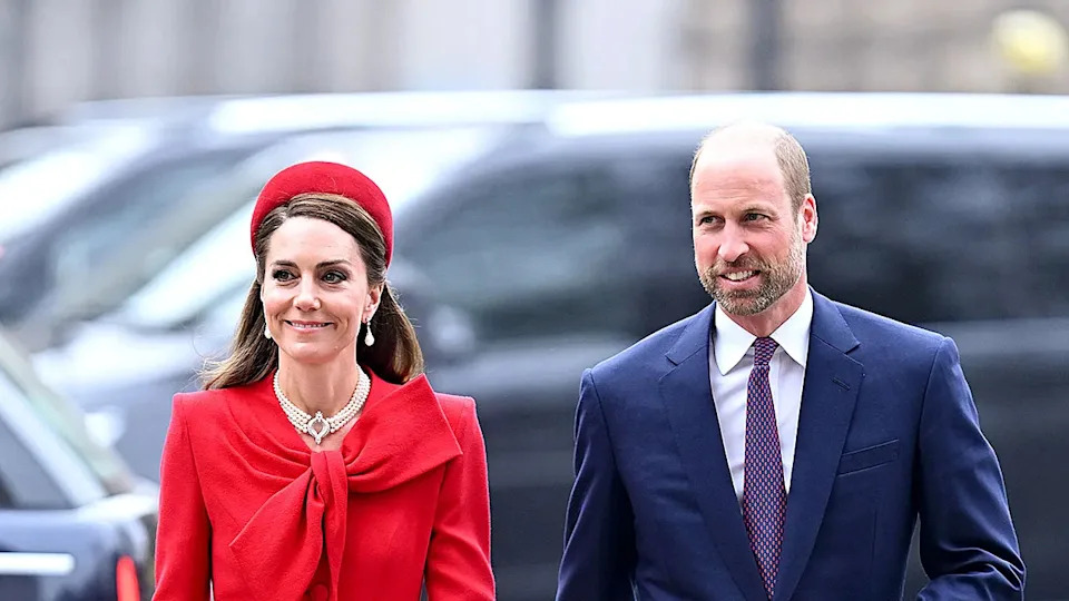 Kate Middleton wearing a red dress and matching hat with pearls walking next to Prince William wearing a blue suit and purple tie.