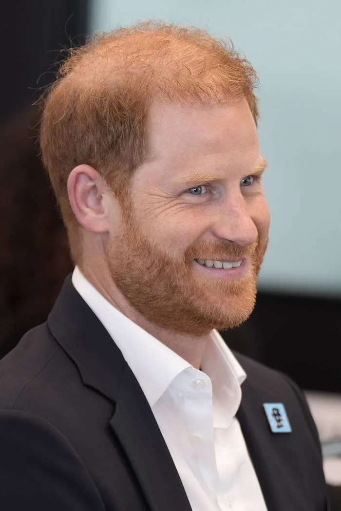 Prince Harry with short red hair and a beard, wearing a suit jacket and white shirt, looks to his left