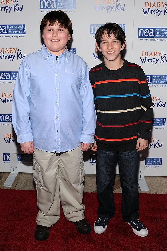 Two young actors stand on a red carpet event for "Diary of a Wimpy Kid," both smiling. One wears a button-up shirt, the other a striped sweater