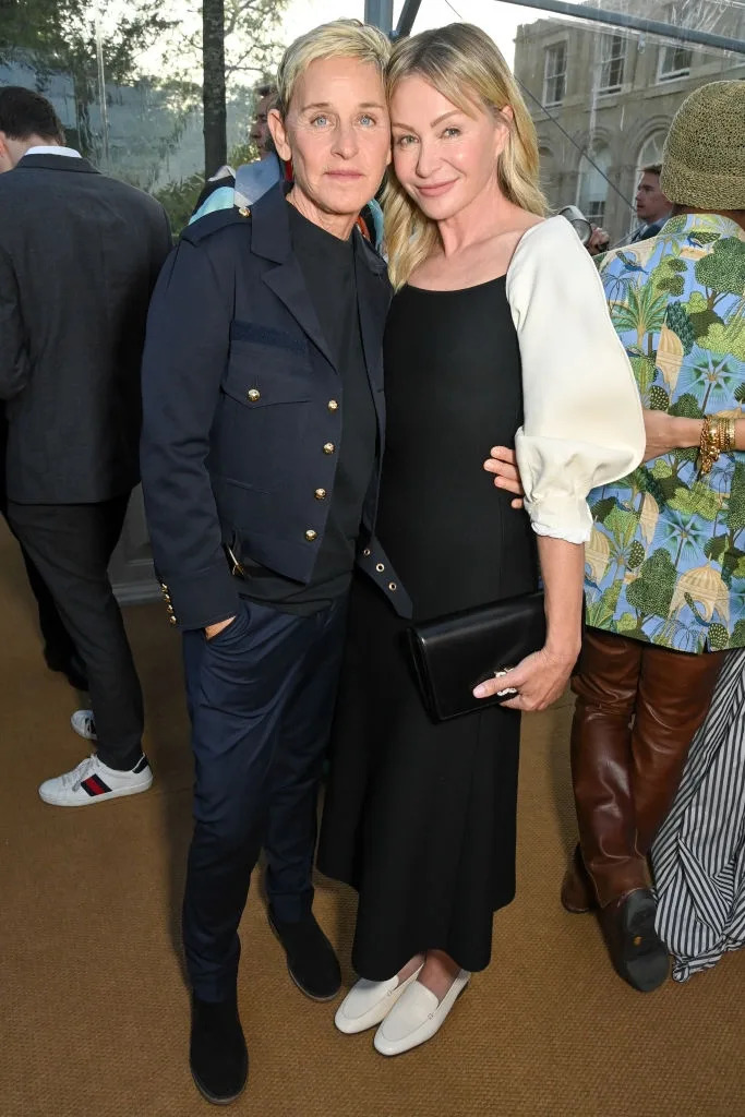 Two people posing at an event. One wears a tailored suit with a military-inspired jacket, the other a long dress with puffed sleeves and holds a clutch