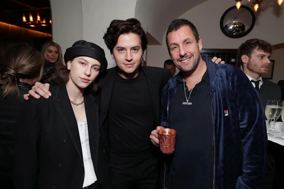 Three individuals in stylish attire at an event, posing together and smiling, with one holding a drink